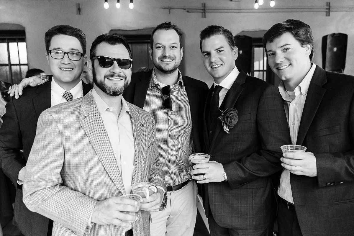 Five men in suits stand together smiling at a social event. They hold drinks, exuding a relaxed and cheerful atmosphere. Black and white photo.