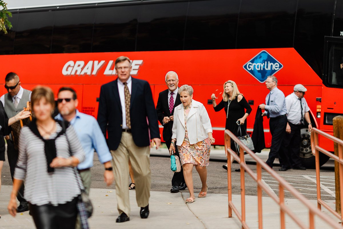 A group of people in formal attire exit a red Gray Line tour bus, smiling and chatting. The scene conveys a cheerful, social atmosphere.