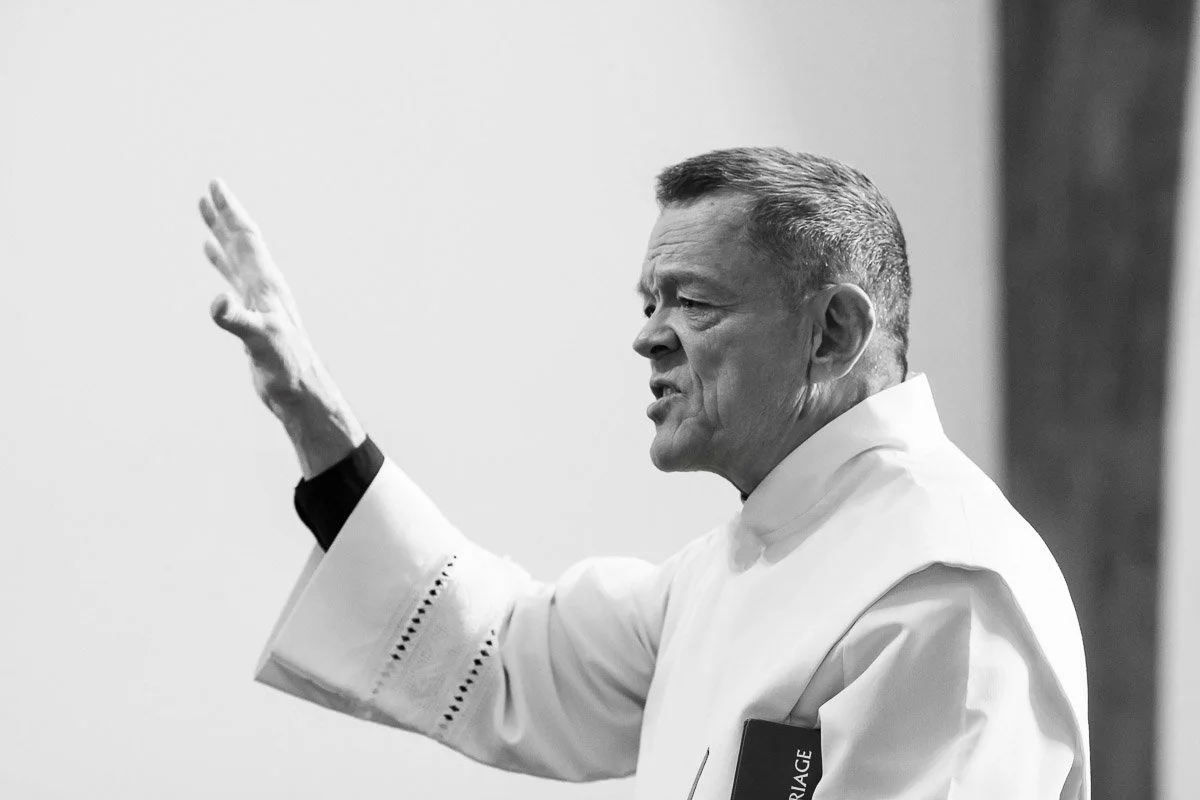 Elderly man in white religious robe gestures with raised hand, speaking passionately. Monochrome setting conveys a solemn and contemplative mood.
