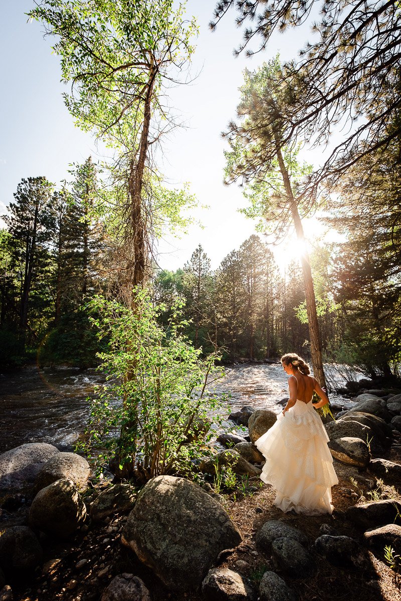 Bride in flowing gown stands by a tranquil river, surrounded by lush trees and boulders, with sunlight filtering through the branches, creating a serene and romantic scene.