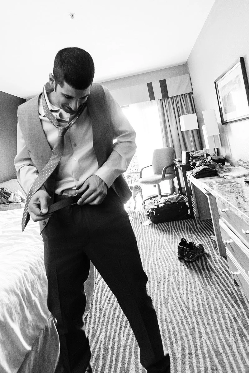 Man adjusts suit while smiling in a hotel room, conveying excitement. Disorganized room with a bed, sneakers, and open suitcase visible. Black and white.