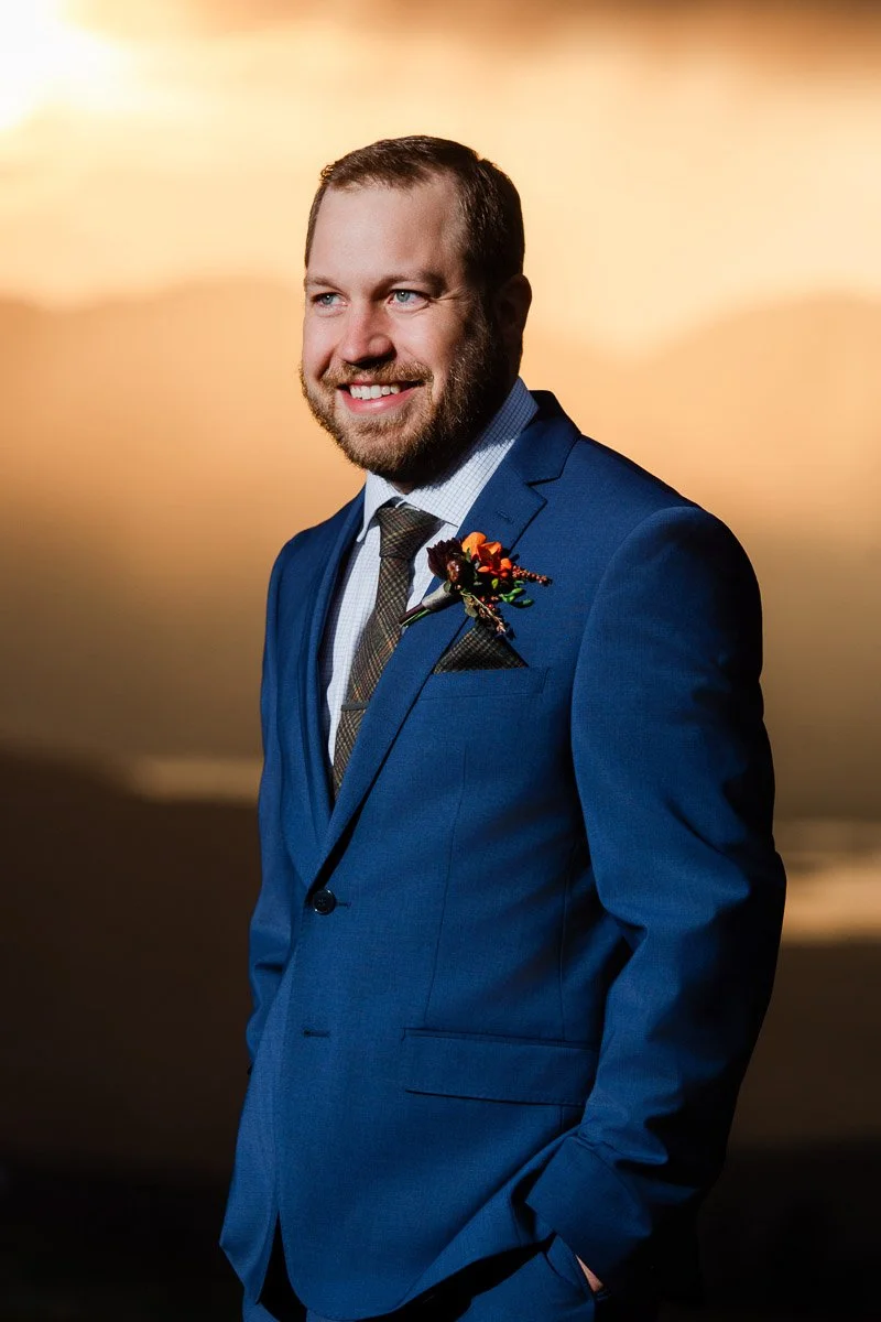 A man in a blue suit smiles warmly against a soft, orange sunset backdrop. He wears a floral boutonniere, exuding a joyful, relaxed mood.