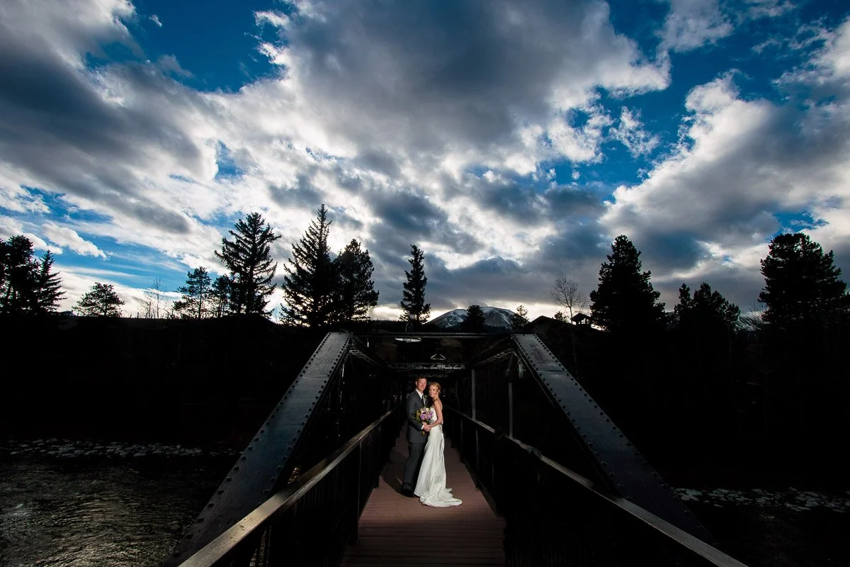 A couple in wedding attire stands on a bridge under a dramatic, cloud-filled sky with silhouetted trees. The scene conveys romance and serenity during a Silverthorne Pavilion wedding