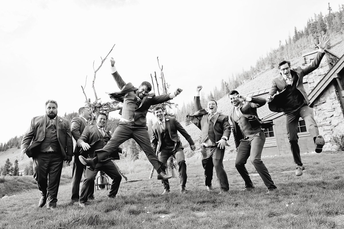 A group of eight men in suits joyfully jumping in the air on a grassy field, with a rustic house and trees in the background. The mood is celebratory and energetic.