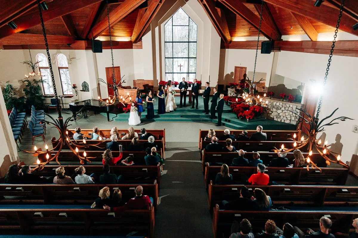 A wedding ceremony in a wooden-ceiling chapel with a large window. Guests sit in pews, while the wedding party stands at the altar. Warm, festive atmosphere.