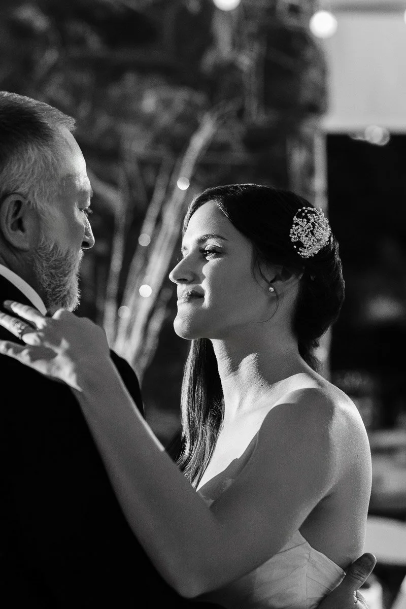 A bride and an older man share a heartfelt dance. She's gazing up with a warm smile, while he looks serenely at her. The setting is intimate and joyful.