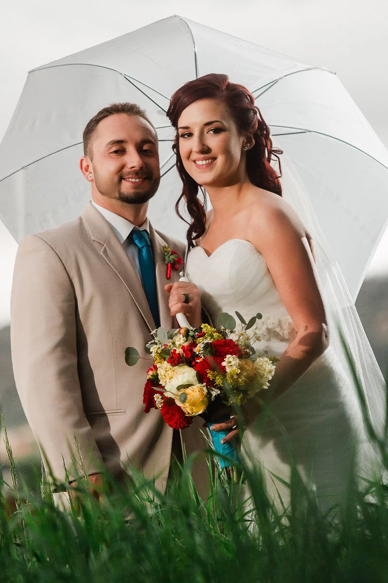 A joyful bride and groom stand closely, holding a vibrant bouquet under a white umbrella. The groom wears a beige suit; the bride is in a white gown.