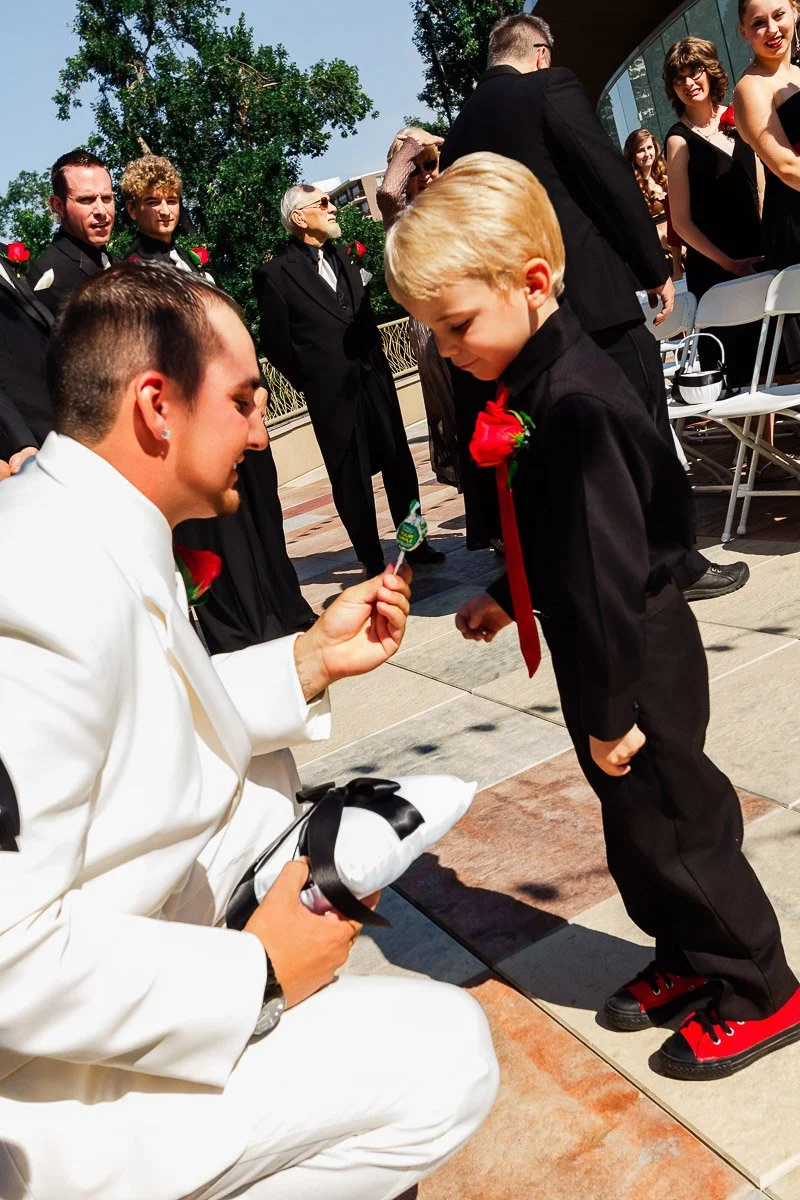 A man in a white suit kneels, offering a lollipop to a young boy in a black suit. Both have red rose boutonnieres. Guests in formal attire observe warmly.