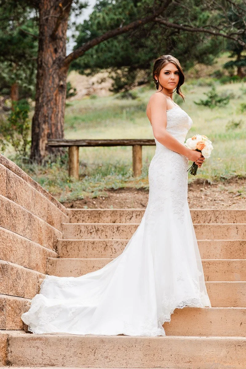 A bride in a strapless white gown stands on the stone steps of Taharaa Mountain Lodge, holding a bouquet of white and peach flowers. She is surrounded by trees and greenery.