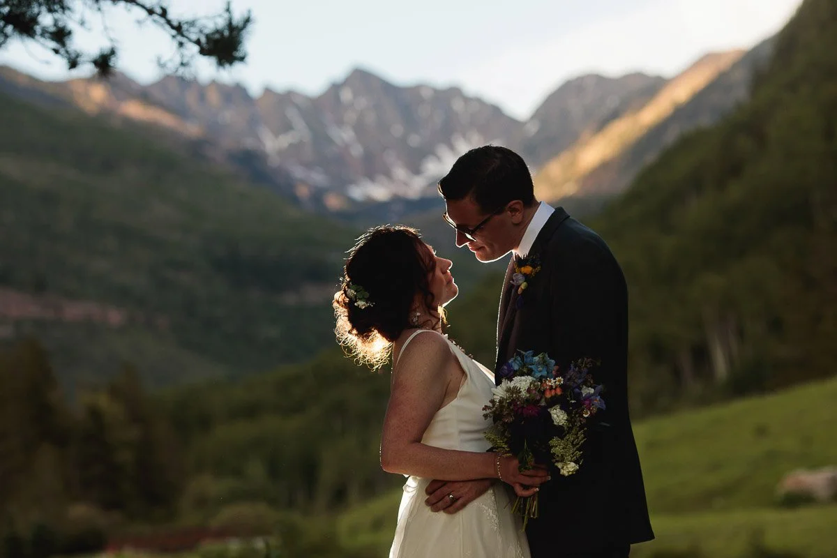 Bride and groom gaze at each other lovingly in a mountainous landscape. The bride holds a colorful bouquet; both are framed by forested hills.