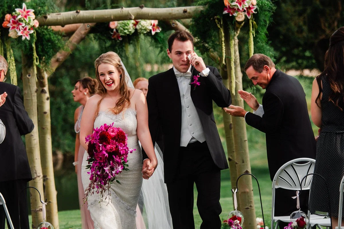 Bride and groom walk down the aisle, hand in hand, under a floral arch. The bride beams joyfully, while the groom wipes a tear, surrounded by clapping guests.