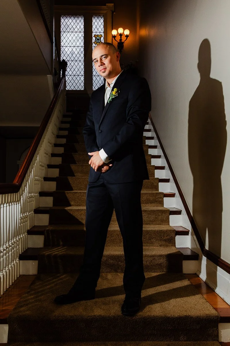 A groom in a suit stands confidently on a carpeted staircase, lit by warm interior lighting. A shadow is cast on the wall, creating a dramatic effect during a Grant Humpreys Mansion wedding in Denver, Colorado.
