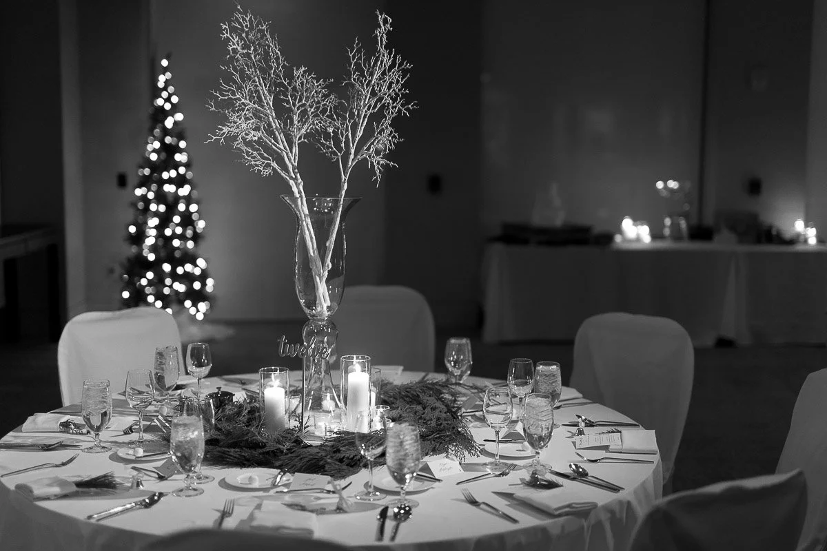 Black and white image of an elegantly set table with a tall vase, branches, and candles. A decorated Christmas tree glows softly in the background.