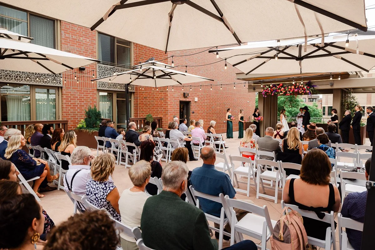 Outdoor Elizabeth Hotel wedding ceremony on a patio. Guests seated on white chairs under large umbrellas watch the couple exchange vows. Festive and romantic atmosphere.