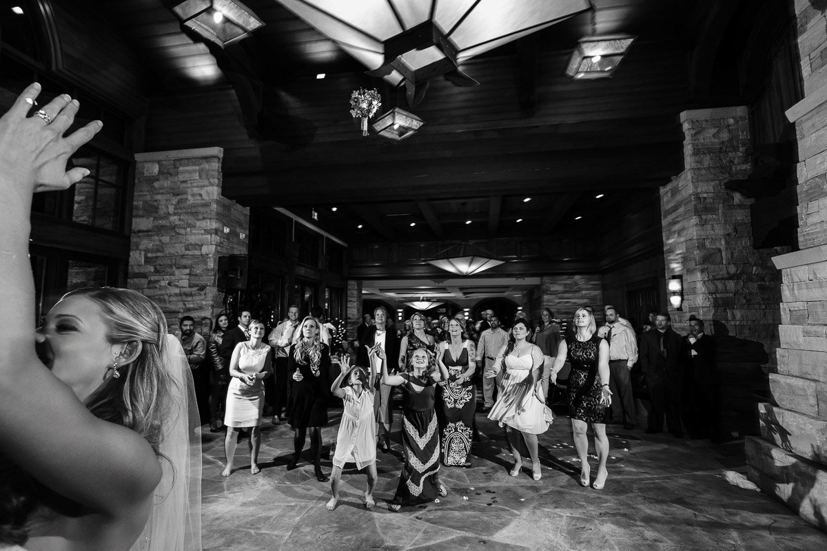 Bride tossing bouquet in elegant venue; group of guests eagerly watching, some reaching up. Joyful and festive atmosphere, captured in black and white.