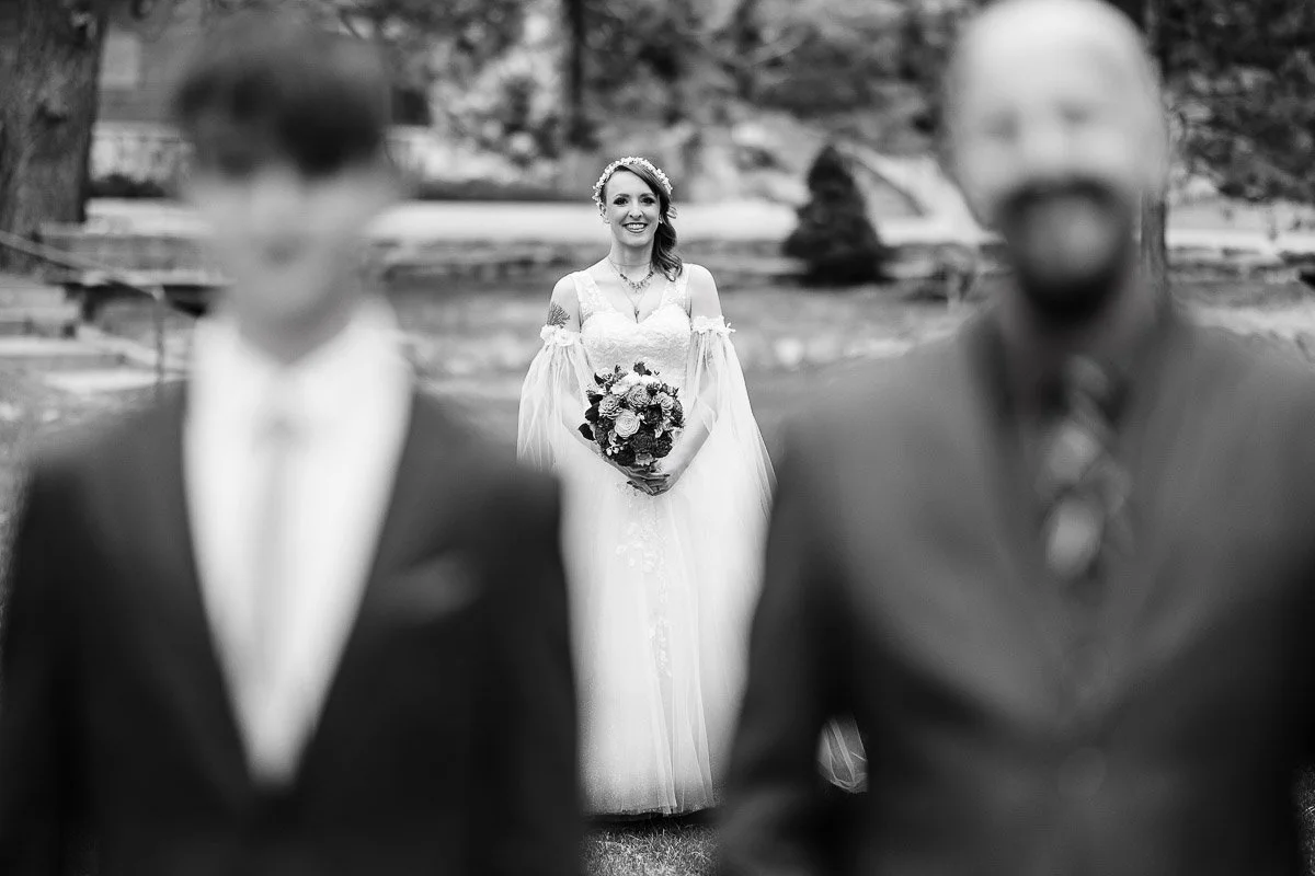 In a black-and-white image, a smiling woman in a wedding dress holds a bouquet, sharply focused between two blurred men in suits in the foreground.