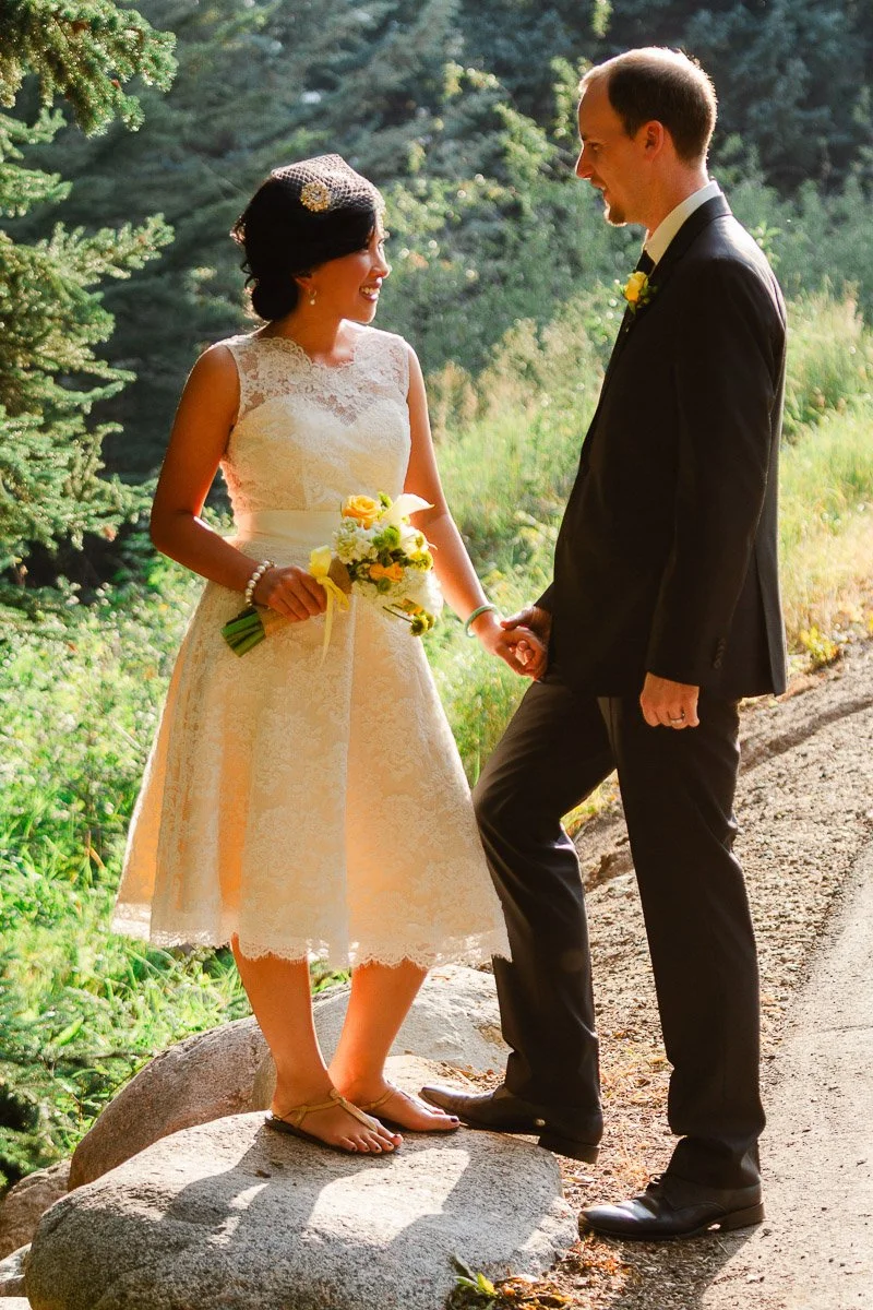 A bride and groom stand holding hands on a sunlit path in a forest. The bride wears a knee-length lace dress and holds a yellow bouquet. The groom is in a suit, smiling warmly.