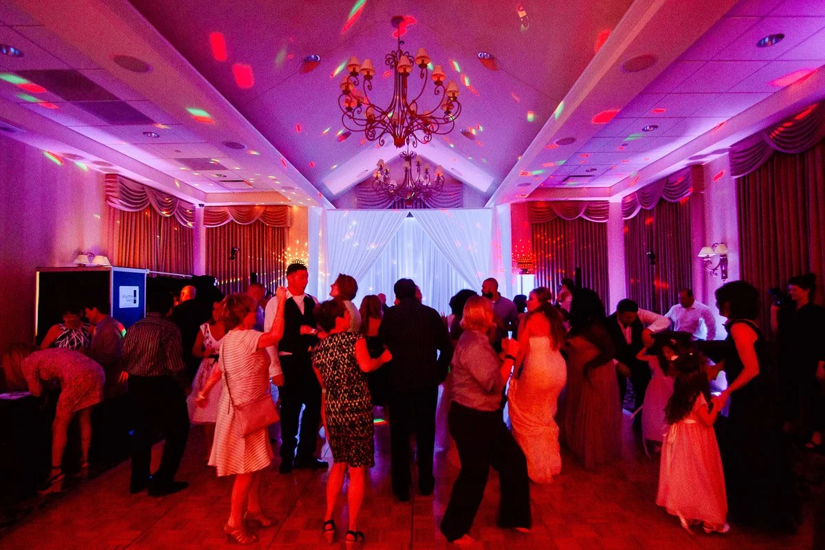A lively party scene in a decorated ballroom, with people dancing under purple and red lights. The atmosphere is joyful and festive.