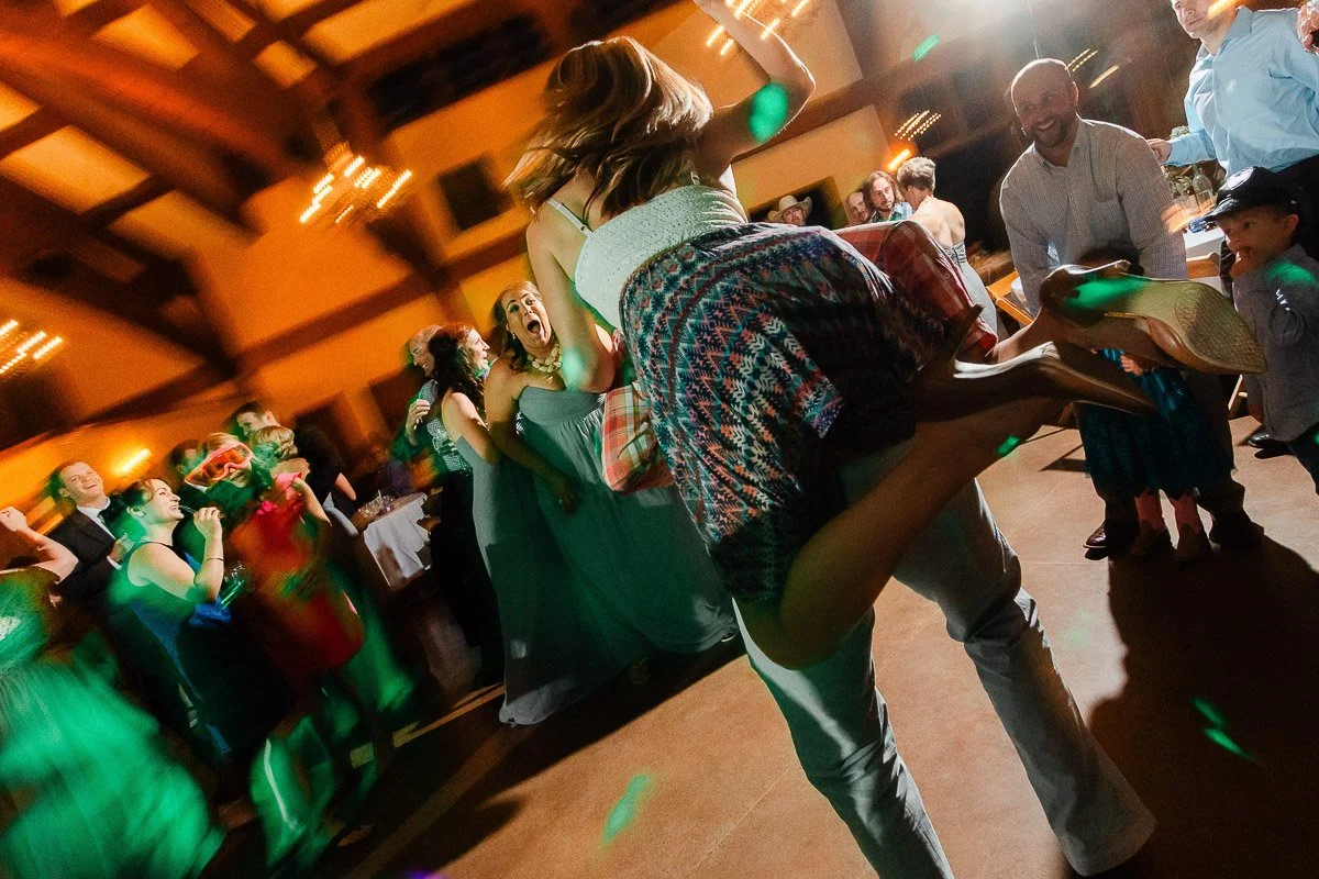 A lively dance floor scene with a joyful woman being lifted mid-air by a man. Guests in formal attire are gathered around, laughing and clapping. Warm lighting and blurred motion convey excitement and celebration.