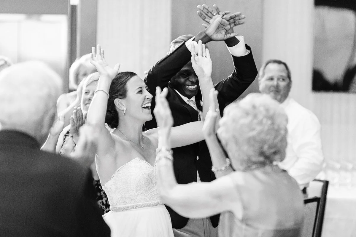 A joyful wedding scene in black and white with a smiling couple dancing among guests. The atmosphere is lively and filled with celebration.