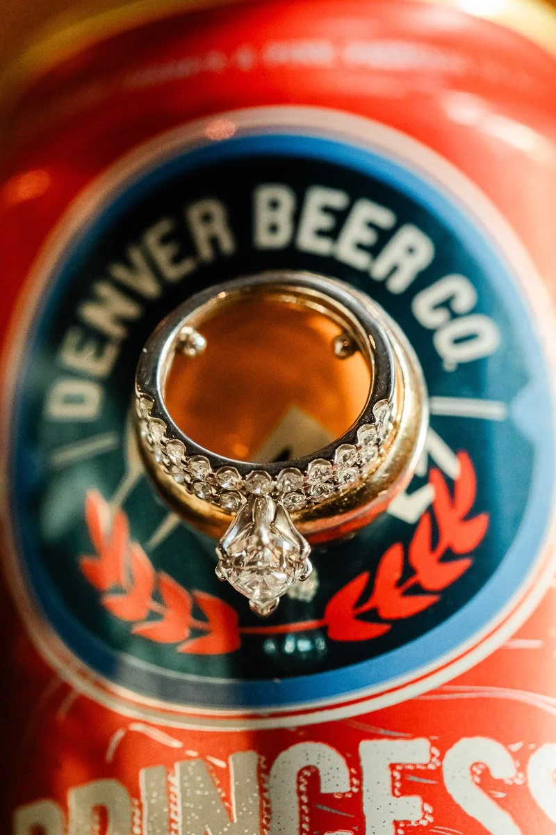 A diamond ring with small gems encircles a beer can's opening, highlighting the "Denver Beer Co" logo with red, blue, and white colors. Playful and unique.