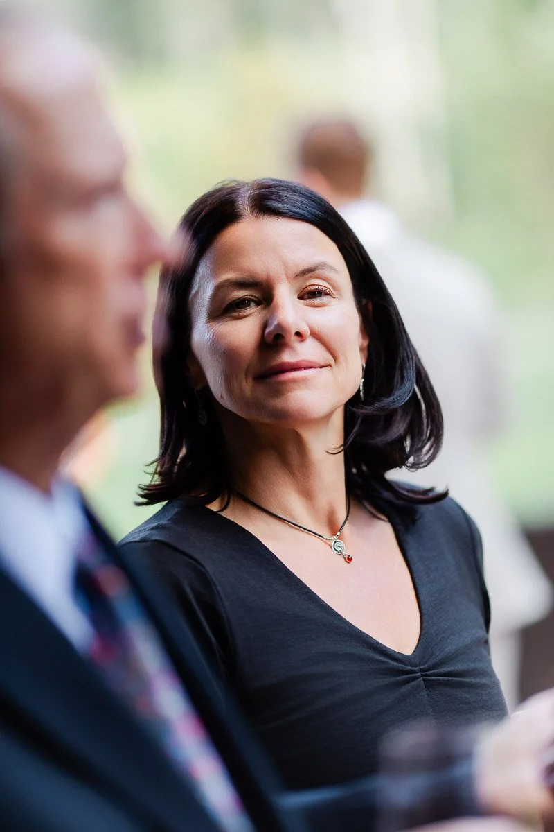 A woman with dark hair and a black top smiles softly while looking at an older man in a suit. The background is blurred, suggesting an outdoor gathering.