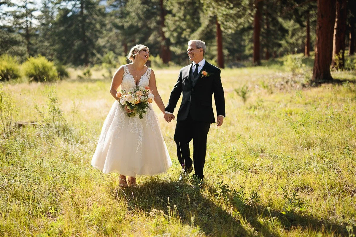 A bride in a white lace dress and floral crown smiles at the groom in a black suit. They walk hand in hand through a sunlit forest meadow during a Della Terra wedding.