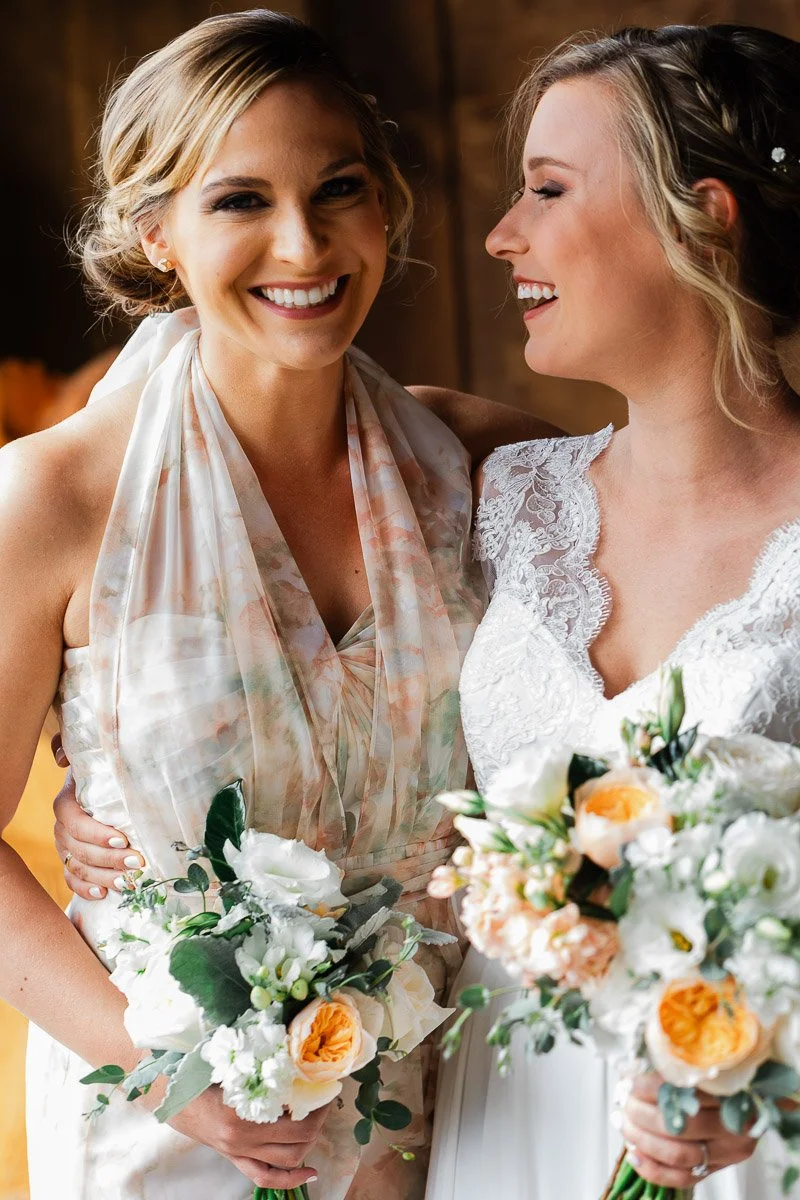 Two smiling women in elegant dresses hold bouquets of white and peach flowers. They stand closely, sharing a joyful moment, conveying warmth and happiness.
