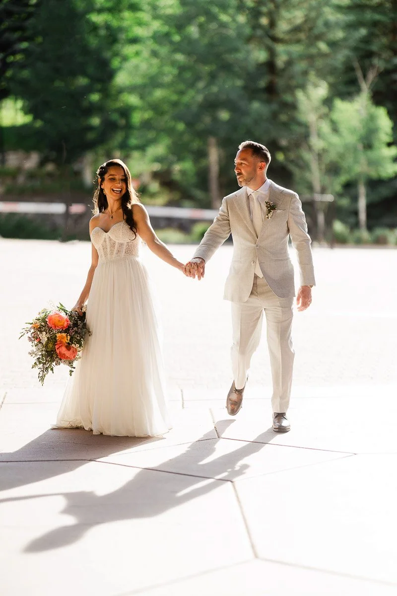 A joyful bride and groom hold hands, walking in sunlight. The bride wears a flowing gown, holding a bouquet of orange and pink flowers. The groom is in a light suit.