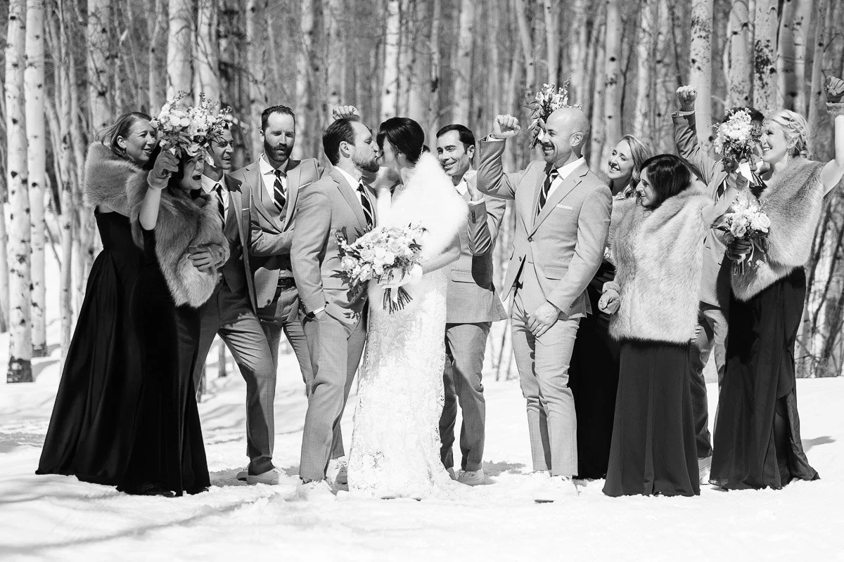 Wedding party in a snowy forest, with the bride and groom kissing in the center. The group is cheerful, wearing elegant attire with fur shawls.