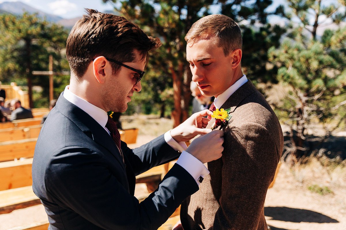 A man in a suit adjusts a boutonniere on another man's jacket at an outdoor wedding. The scene is set against a backdrop of trees and wooden benches, conveying a sense of focus and camaraderie.