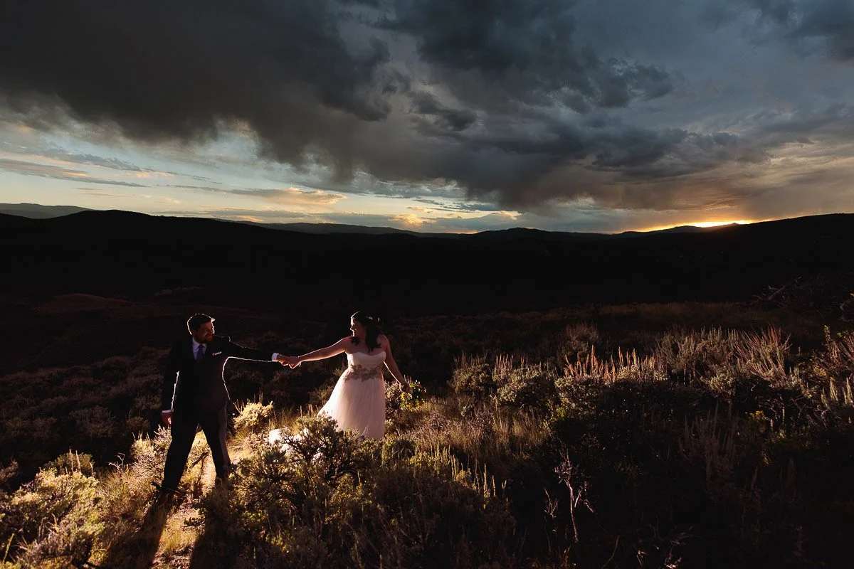 A couple stands holding hands in a serene field at sunset, surrounded by dramatic clouds. Their attire suggests celebration, conveying romance and tranquility during a Westin Riverfront Resort wedding.