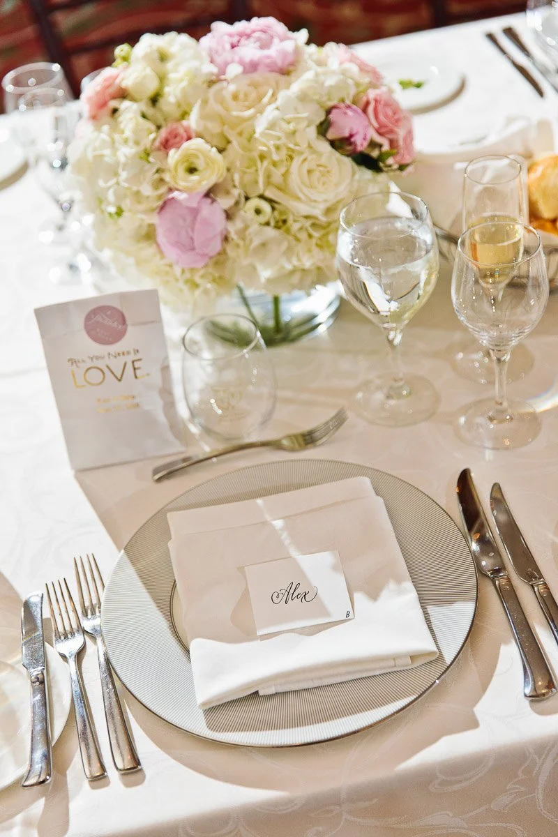 Elegant table setting with a white and silver theme, featuring a name card reading "Alex." A floral centerpiece of pink and white roses adds charm.