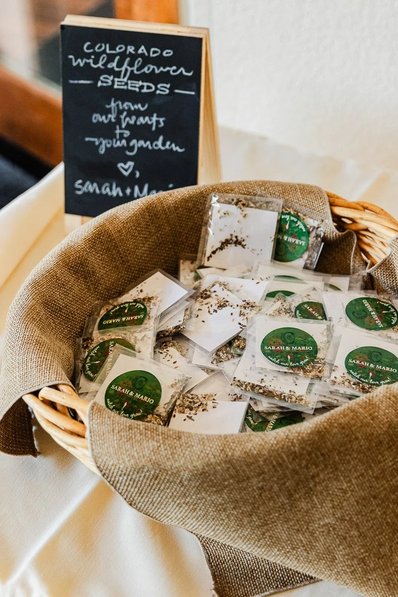 A basket with burlap lining contains packets of Colorado wildflower seeds. A chalkboard sign nearby reads “from our hearts to your garden.” The ambiance is rustic and warm.