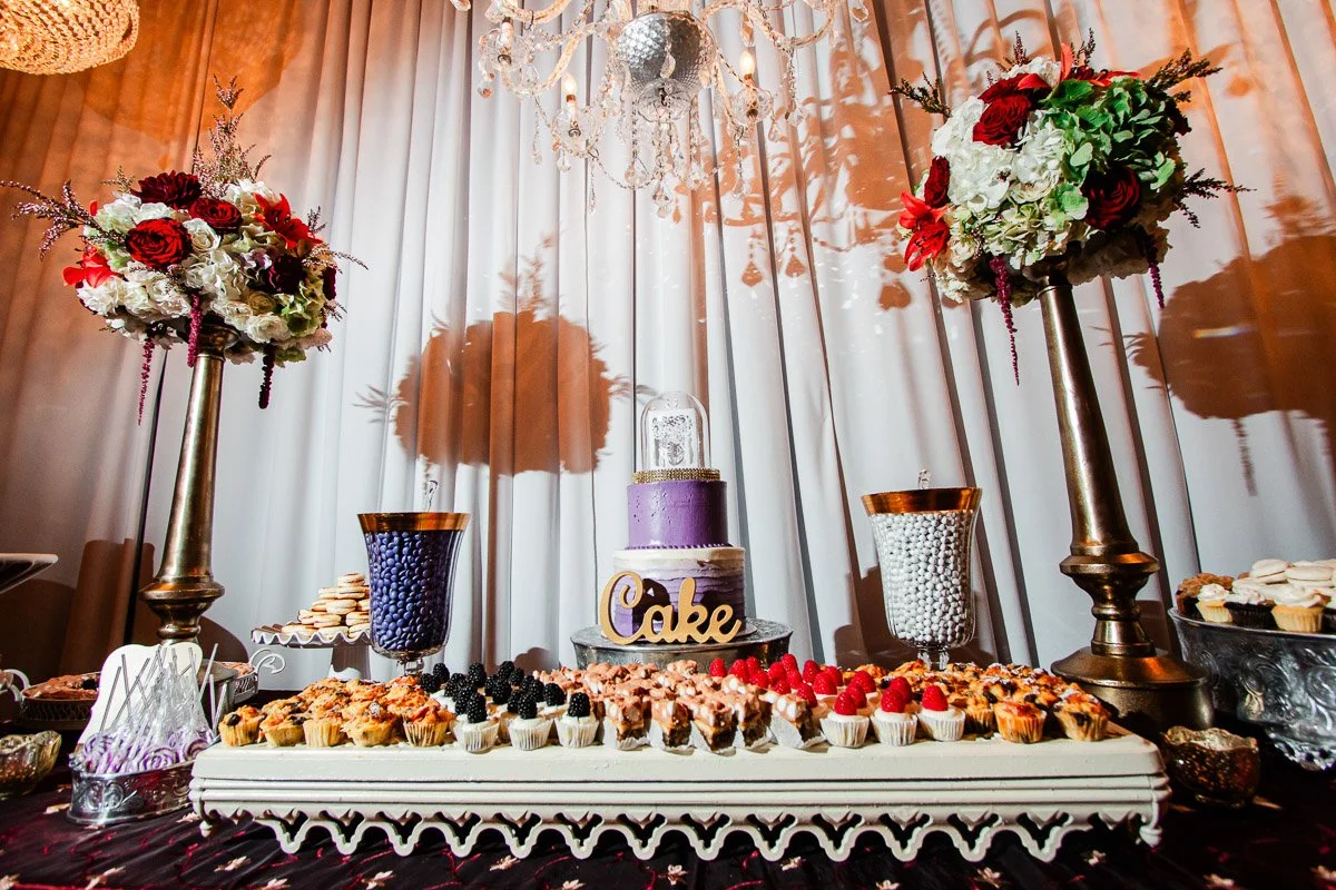 Elegant dessert table with a purple cake, assorted pastries, and vibrant flower arrangements under a chandelier. Luxurious and festive atmosphere.