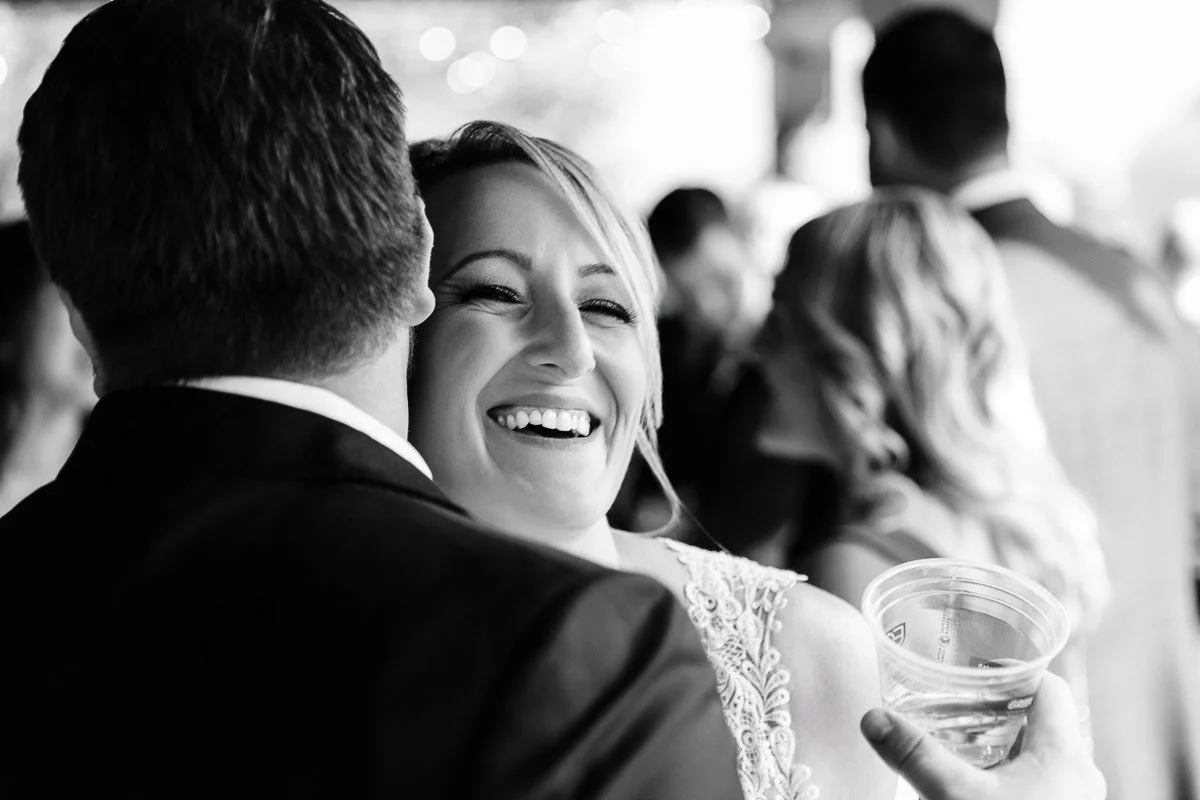 A black-and-white photo of a woman joyfully laughing while hugging a man. She holds a plastic cup, and a crowd mingles in the blurred background. Emotions convey happiness and celebration.