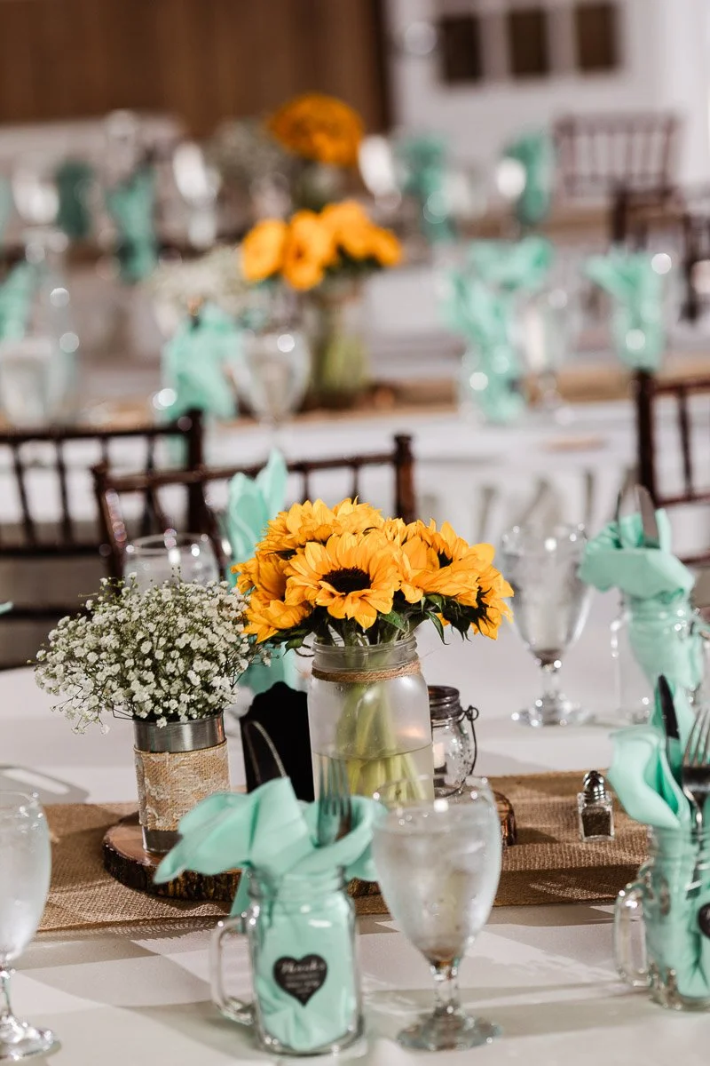 Elegant table setting with vibrant yellow sunflowers in mason jars, teal napkins, and rustic burlap runner. Glasses and silverware add a refined touch.