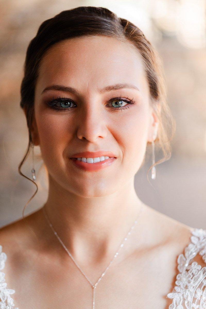 A woman in a white lace dress smiles softly, with subtle makeup and delicate jewelry. Soft lighting creates a warm, serene ambiance.