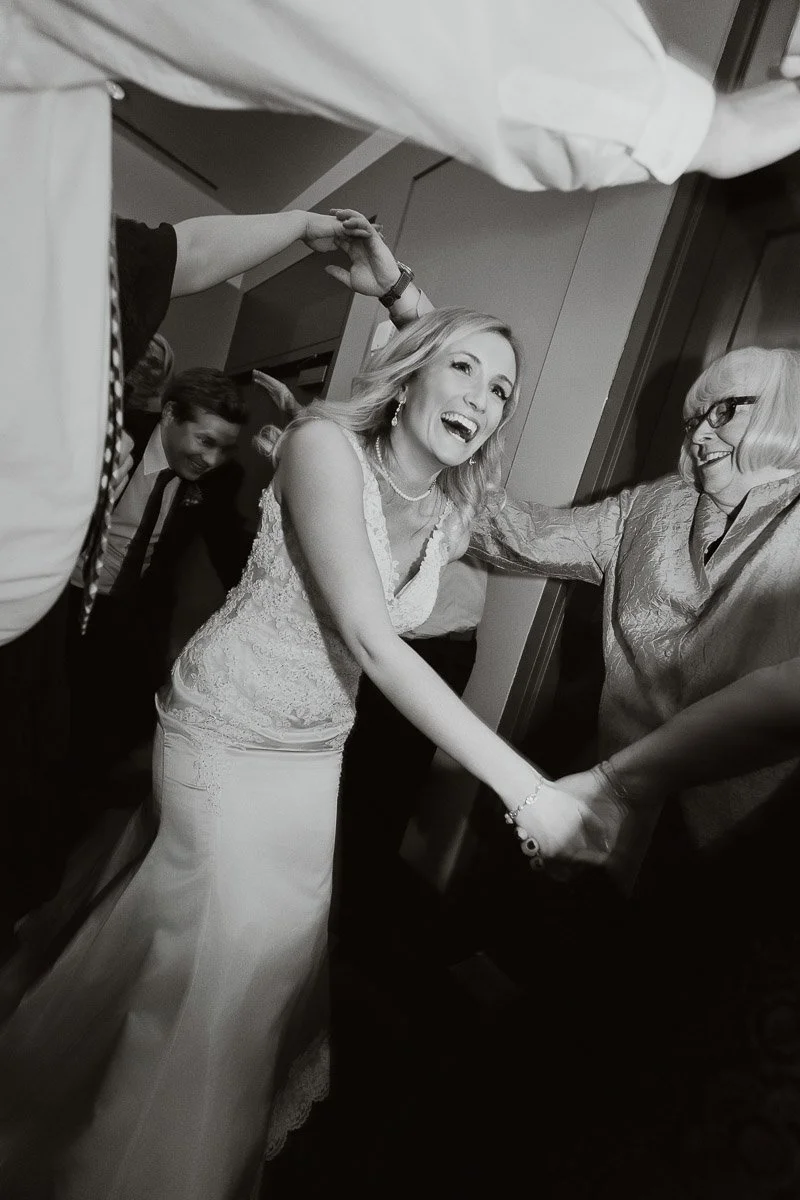 A joyful bride in a lace gown smiles brightly while dancing under raised hands at a celebration. The scene conveys excitement and happiness. Black and white photo.