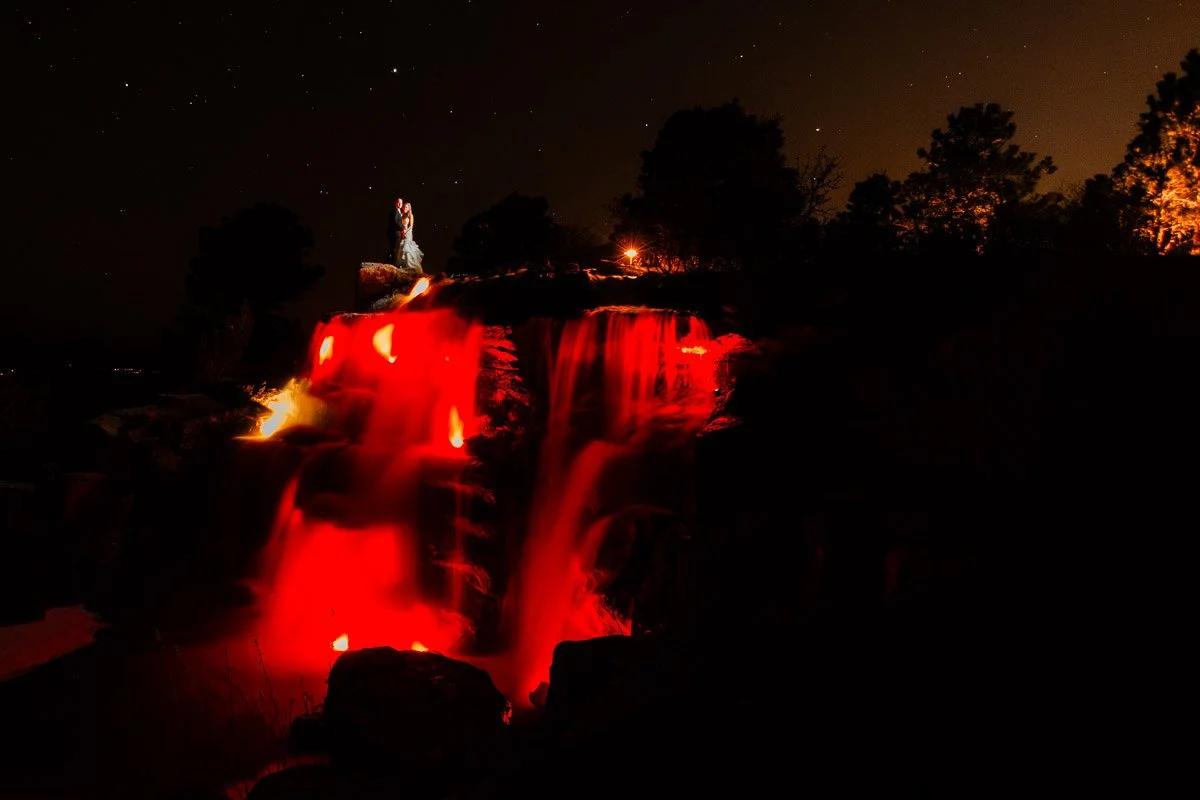 A couple embraces atop a waterfall under a starry sky. The cascading water glows red, creating a dramatic and romantic atmosphere at a Sanctuary Golf Course wedding in Sedalia, Colorado.