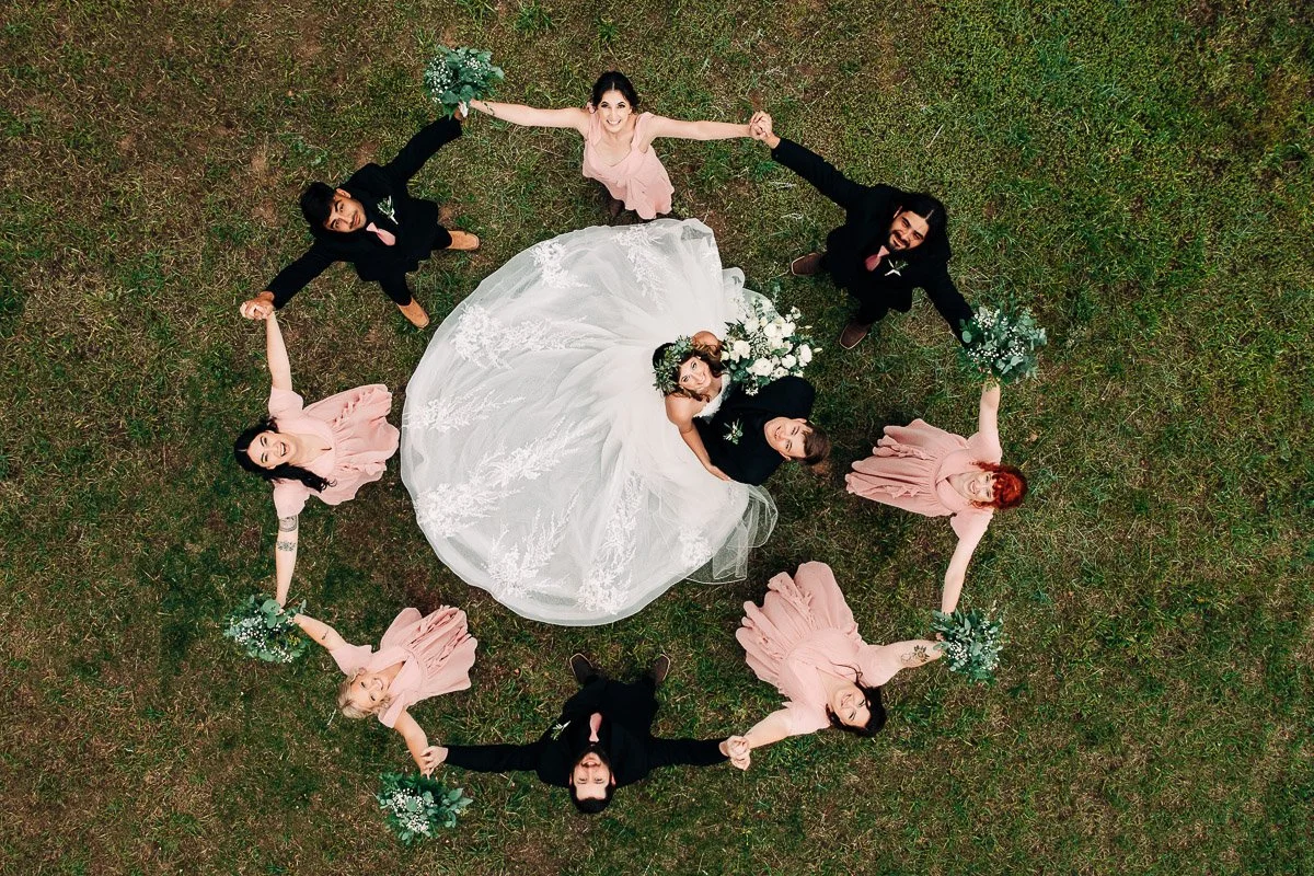 Wedding party forms a joyful circle on grass, viewed from above. Bride in center, surrounded by bridesmaids in pink and groomsmen in black, holding hands captured by Colorado wedding photographer tomKphoto
