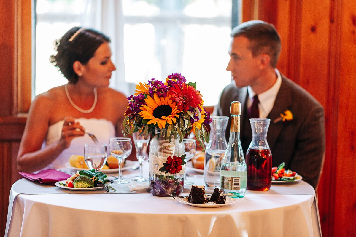 A couple in formal attire sits at a table with vibrant flowers and wine. The setting is warmly lit, suggesting a celebratory, intimate atmosphere.