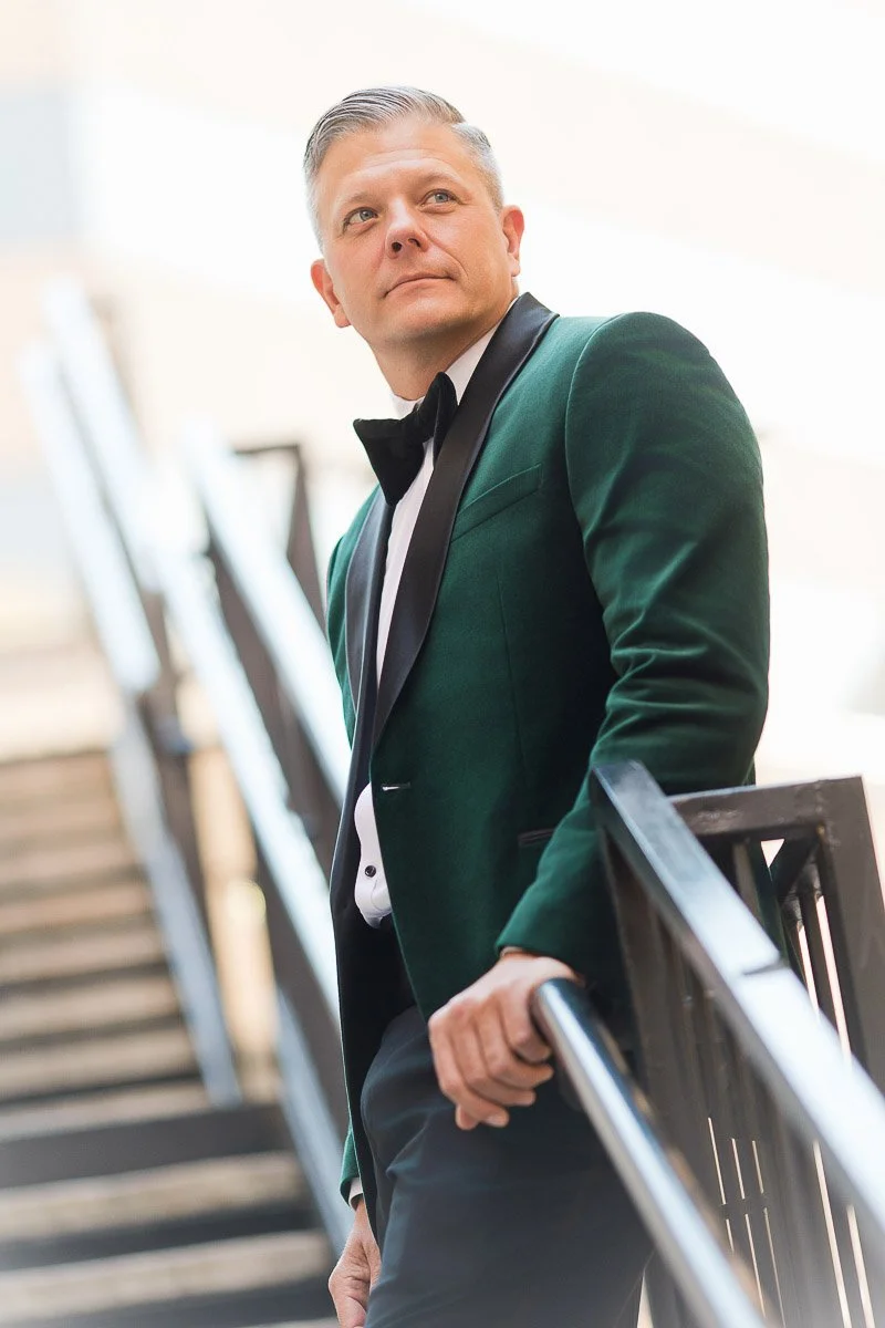 Man in a green velvet jacket and bow tie leans on a railing, gazing thoughtfully upward. The background shows blurred stairs and soft lighting.