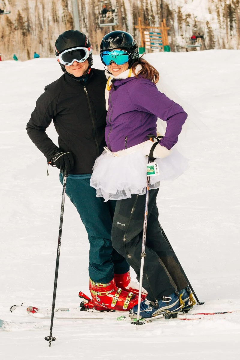 A couple in ski gear embraces on a snowy slope. The woman wears a purple jacket with a tutu, creating a playful, lighthearted atmosphere.