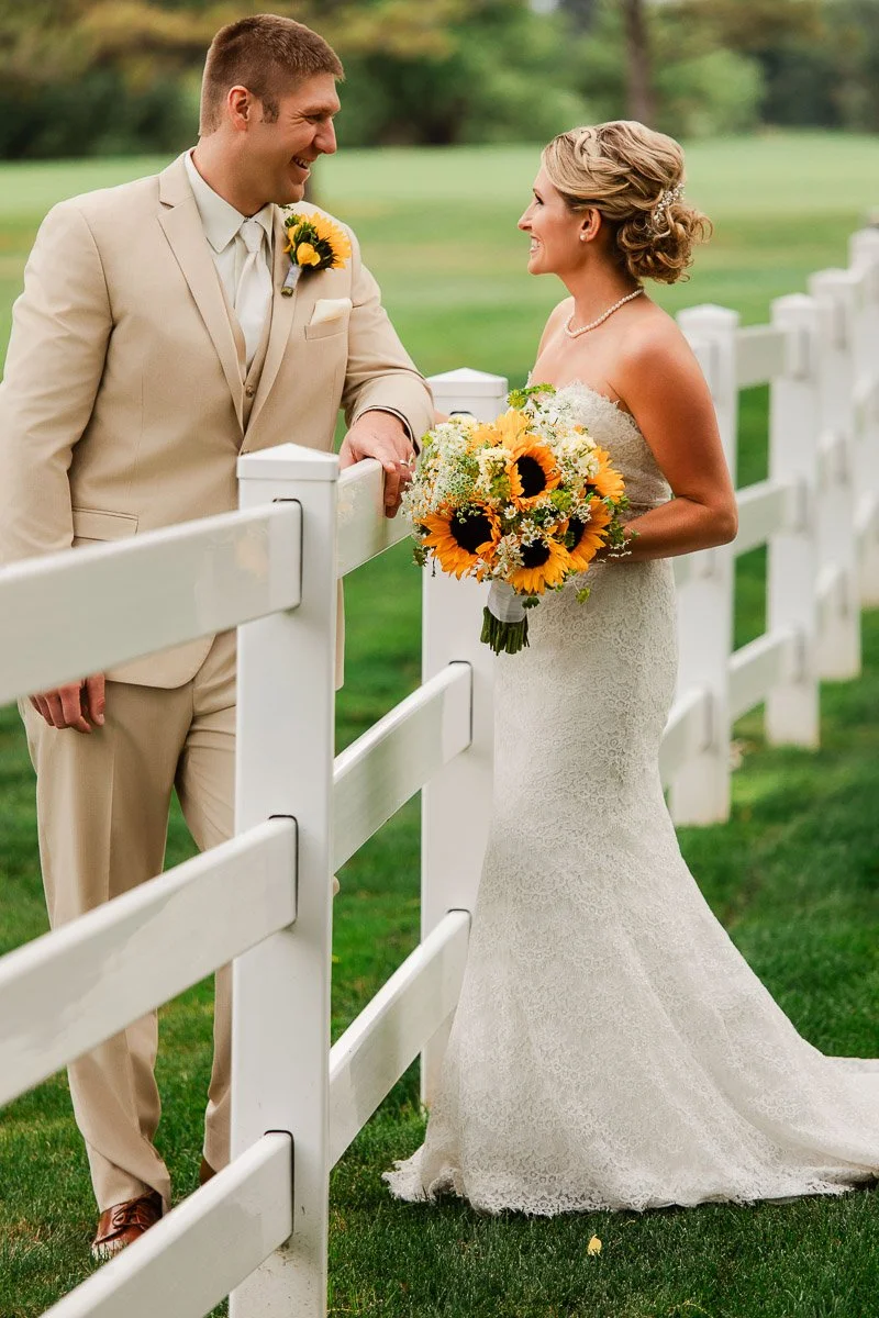 Bride in a lace gown holding sunflower bouquet smiles at groom in beige suit by a white fence on a green lawn, conveying joy and romance during a Barn at Raccoon Creek wedding in Littleton, Colorado