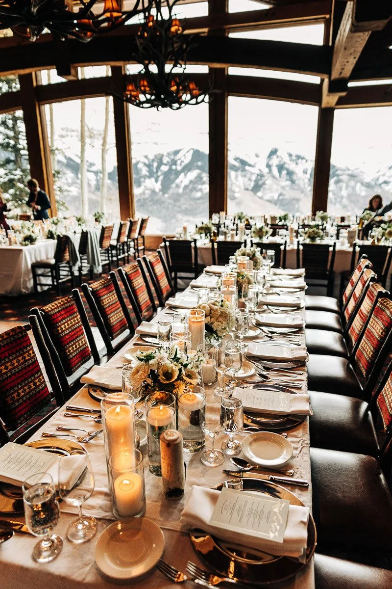 Elegant dining table set for a formal event, featuring candles, flower arrangements, and menus. Large windows reveal a snowy mountain view. Cozy ambiance by Beaver Creek wedding photographer tomKphoto