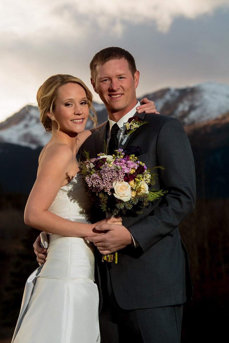 A happy couple poses in wedding attire, bride in white dress, groom in dark suit, holding a colorful bouquet, with majestic mountains in the background.