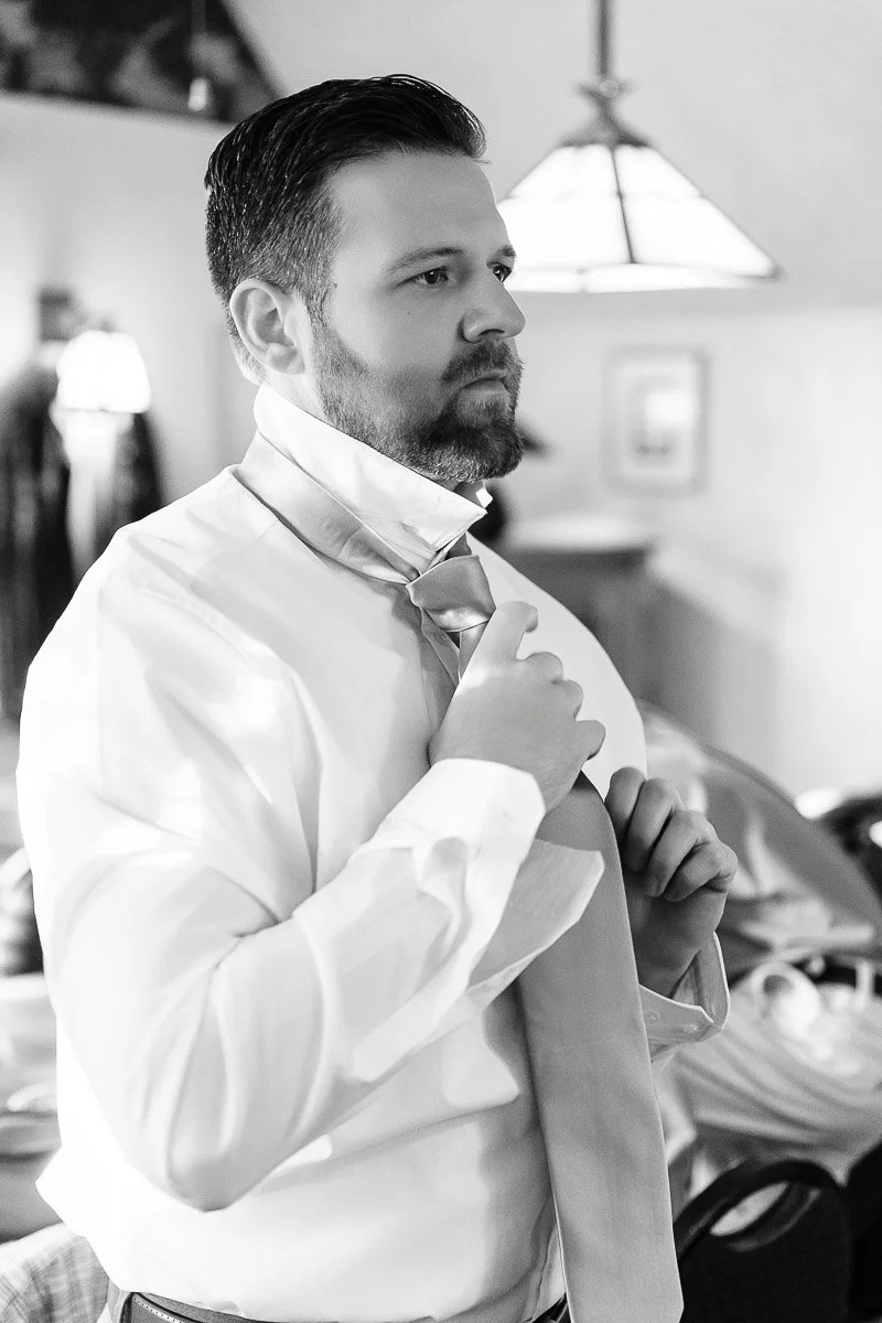 A man in a white shirt adjusts his tie with a focused expression in a softly lit room. The setting is relaxed, with blurred furniture in the background.