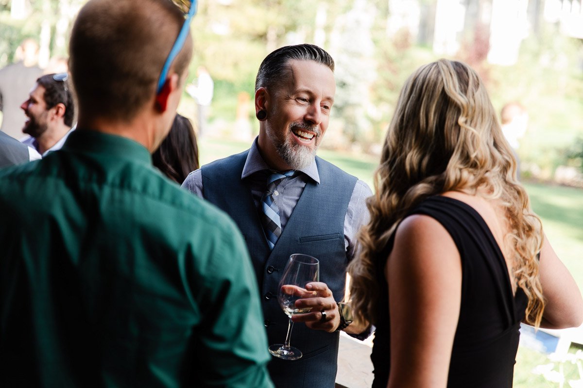 A man with a beard, wearing a vest, smiles while holding a wine glass and talking to two people outdoors at a lively gathering.