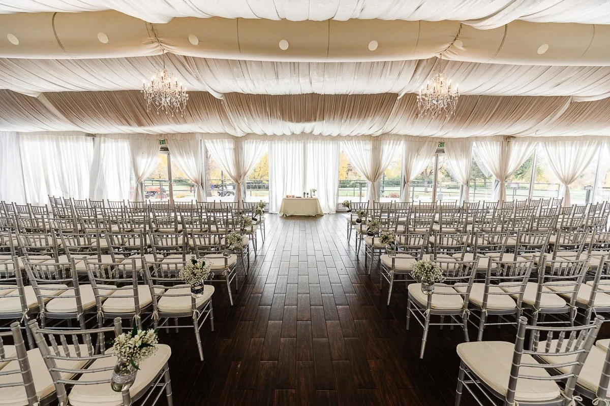 Elegant wedding venue with rows of silver chairs, floral aisle decorations, and a wooden floor. White draped ceiling and chandeliers create a romantic atmosphere.